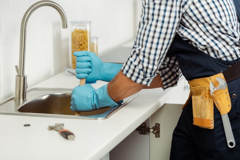 Cropped view of plumber in tool belt and rubber gloves cleaning blockage of kitchen sink