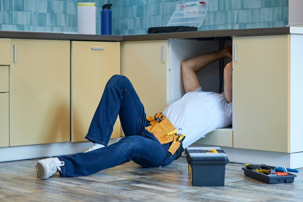 Efficient work. Professional plumber wearing tool belt fixing sink pipe indoors. Tools and equipment on the floor in the kitchen. Renovation and plumbing concept. Horizontal shot