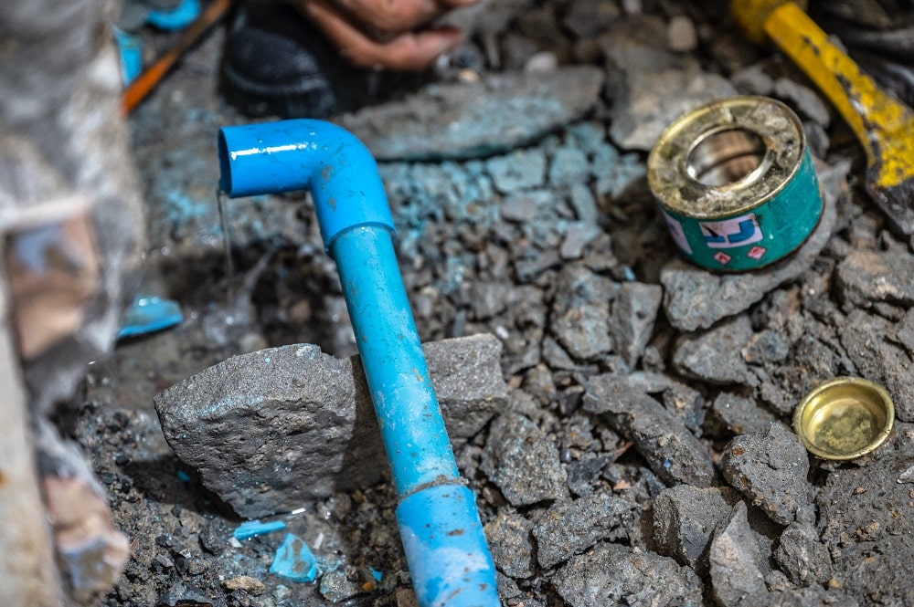 Close-up of a leaking blue PVC pipe surrounded by debris and tools, with water dripping from the joint and a container of adhesive nearby—illustrating why pipe burst issues often occur at weak connections.