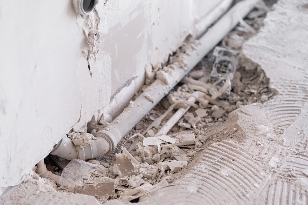 Exposed pipes and broken debris along a damaged wall and floor suggest possible Slab Leaks Repair, with visible dust and construction material scattered around.