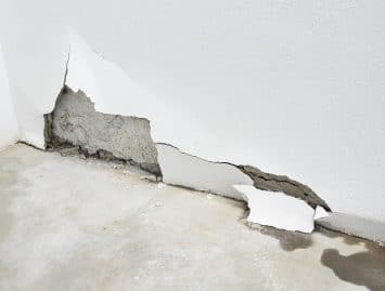 A white wall with a large section of plaster broken off reveals the underlying concrete and water stains on the floor, suggesting potential slab leaks repair may be needed.