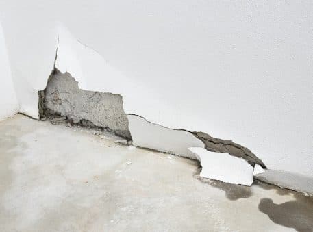 A white wall with a large section of plaster broken off reveals the underlying concrete and water stains on the floor, suggesting potential slab leaks repair may be needed.