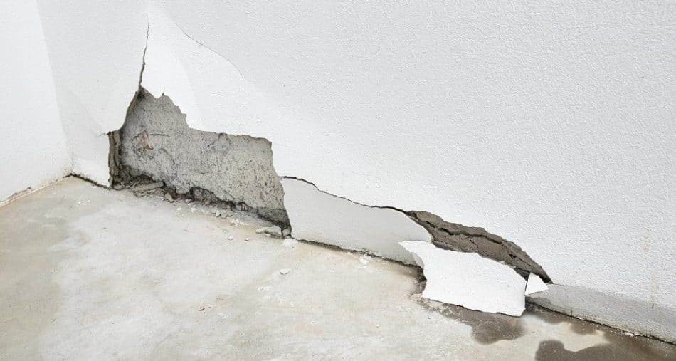 A white wall with a large section of plaster broken off reveals the underlying concrete and water stains on the floor, suggesting potential slab leaks repair may be needed.