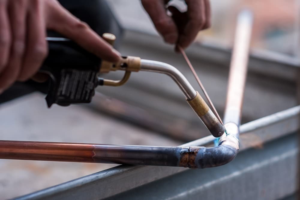 A person uses a blowtorch to solder a copper pipe joint, holding a metal rod to apply the solder—an essential step for preventing leaks and understanding why pipe burst issues may occur.