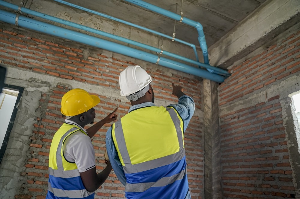 Two workers wearing safety vests and hard hats inspect blue piping during a plumbing renovation in the ceiling of a brick-walled building under construction.