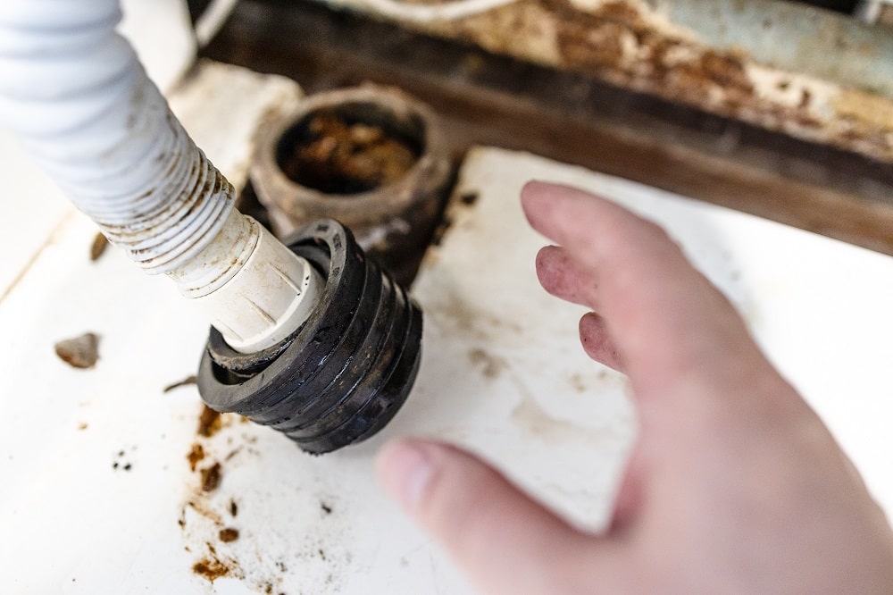 A hand reaches toward a disconnected, dirty plastic pipe fitting on a white surface, with visible grime and rust nearby—highlighting the need for professional drain cleaning.