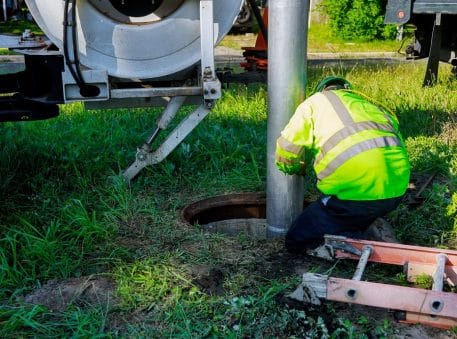 A worker in a high-visibility jacket and helmet operates equipment at an open manhole, providing Sewer Inspection Services in New Orleans, on a grassy area beside a truck and an orange ladder.