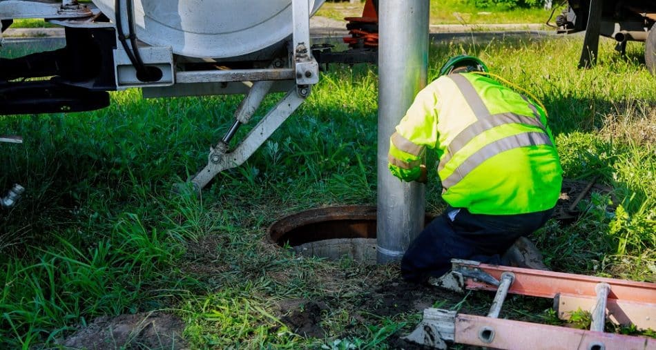A worker in a high-visibility jacket and helmet operates equipment at an open manhole, providing Sewer Inspection Services in New Orleans, on a grassy area beside a truck and an orange ladder.