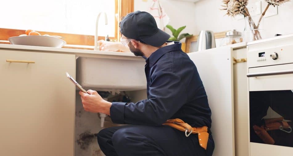 A plumber wearing a navy uniform and cap inspects the pipes under a kitchen sink while holding a clipboard, ensuring everything is ready for professional drain cleaning.