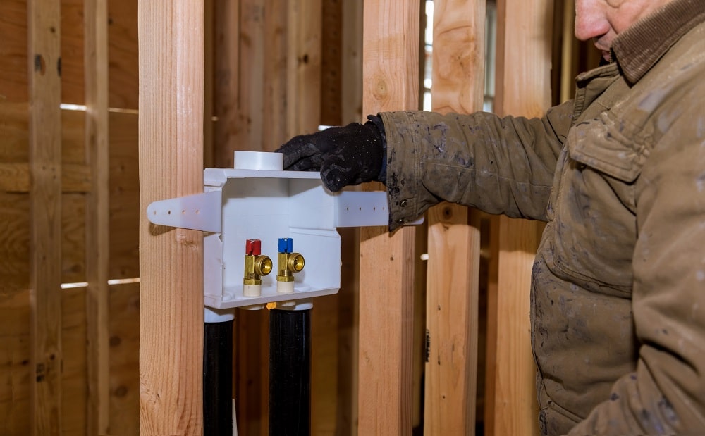 A person in a brown jacket installs or inspects a washing machine outlet box with hot and cold water valves on wooden framing inside a building during a plumbing renovation.
