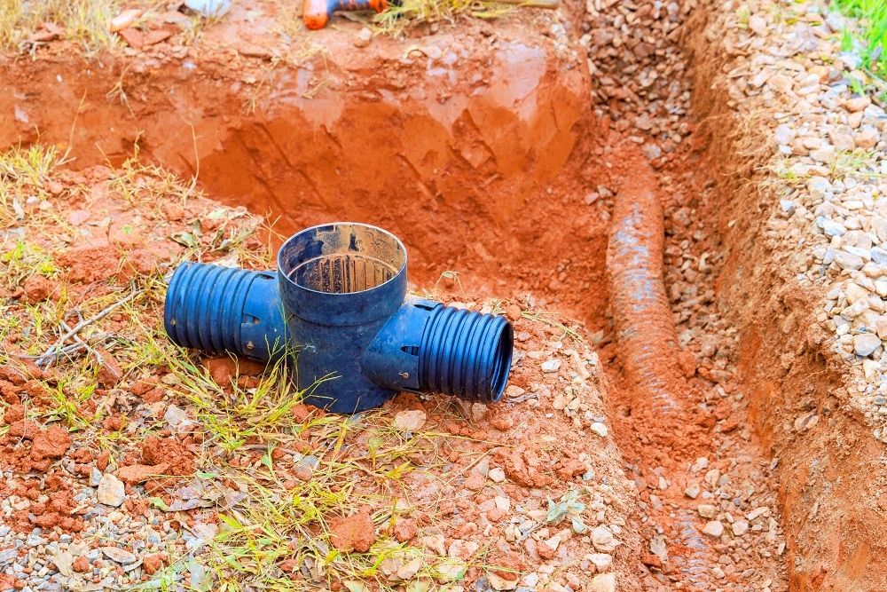 A large plastic pipe fitting lies on red soil next to a trench containing a partially buried corrugated pipe, highlighting the benefits of regular sewer inspections for maintaining underground infrastructure.