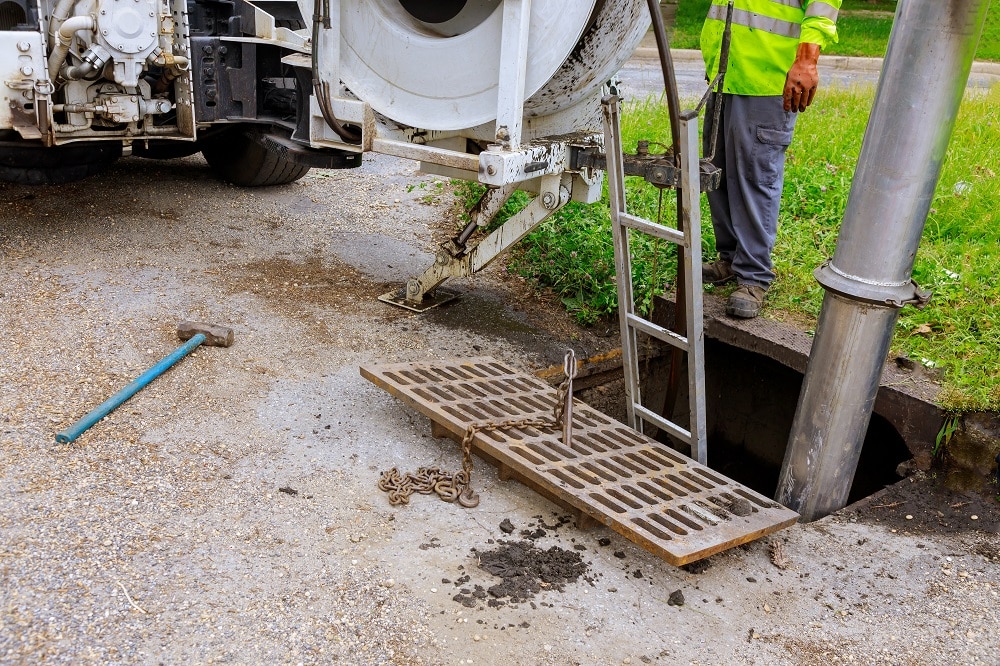 A worker stands by an open storm drain as a metal grate is lifted with equipment attached to a utility truck, illustrating the benefits of regular sewer inspections; tools and chains are scattered on the ground.
