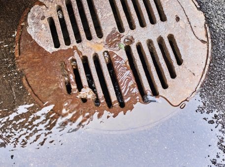 A close-up of a rusty metal storm drain cover with water flowing into it, surrounded by wet pavement—a reminder of the benefits of regular sewer inspections for preventing blockages and maintaining smooth drainage.